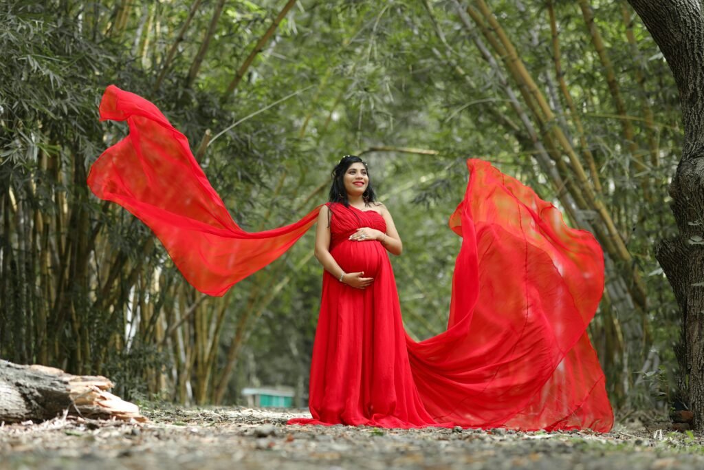 Pregnant woman in a flowing red dress poses gracefully in a lush Indian forest.