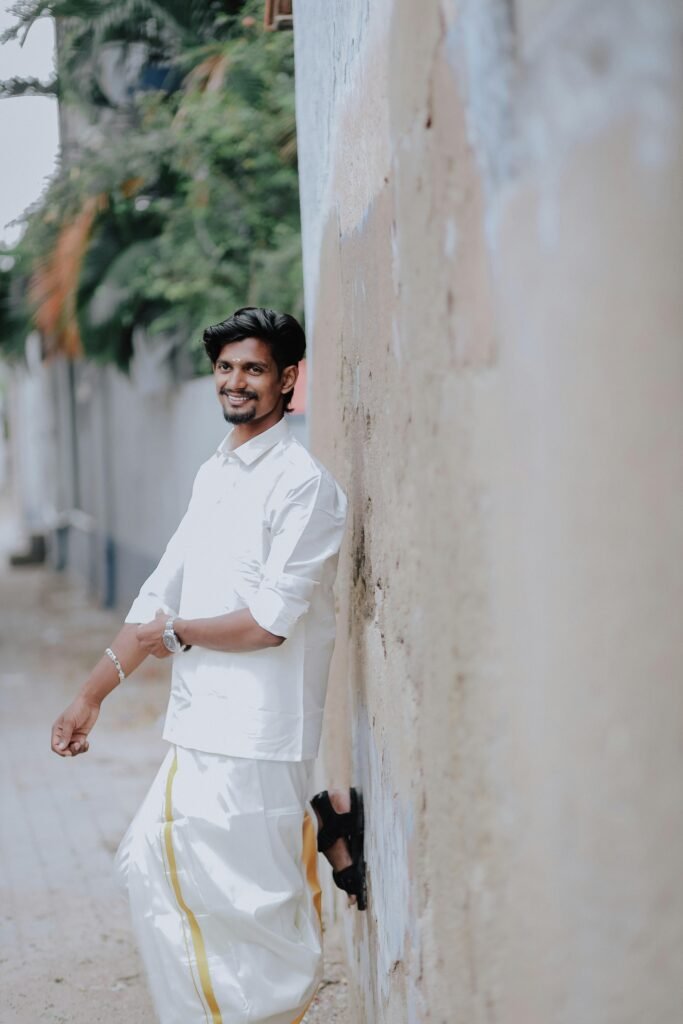 A smiling Indian man wearing traditional attire leaning against a wall outdoors.